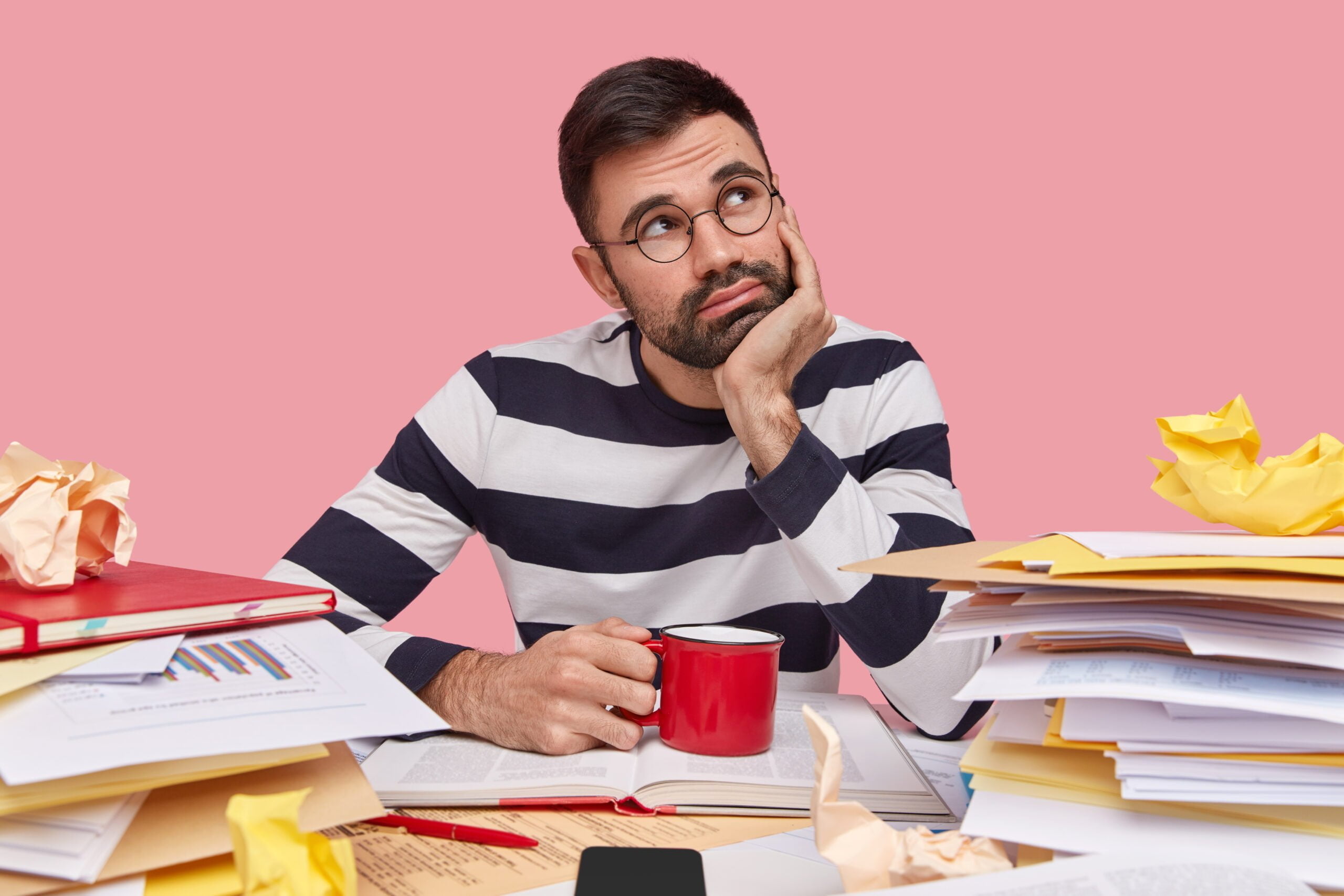 thoughtful young man has contemplative expression holds hand chin wears striped jumper drinks fresh beverage surrounded with pile textbooks scaled