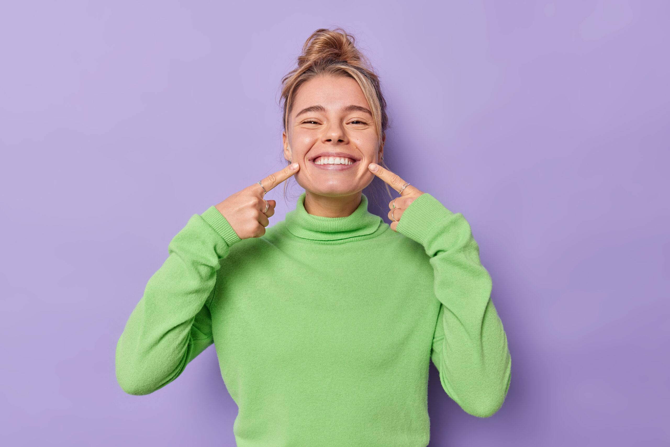 beautiful happy fair haired european woman points index fingers mouth forces cheerful smile shows perfect white teeth wears green jumper being good mood isolated purple background scaled